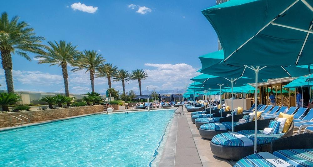 Trump International Hotel Лас Вегас Екстериор снимка The photo depicts a swimming pool scene on a sunny day. On one side of the pool, there are several lounge chairs with blue-striped cushions, each placed beneath large teal umbrellas for shade. Palm trees are visible in the background, providing a tro