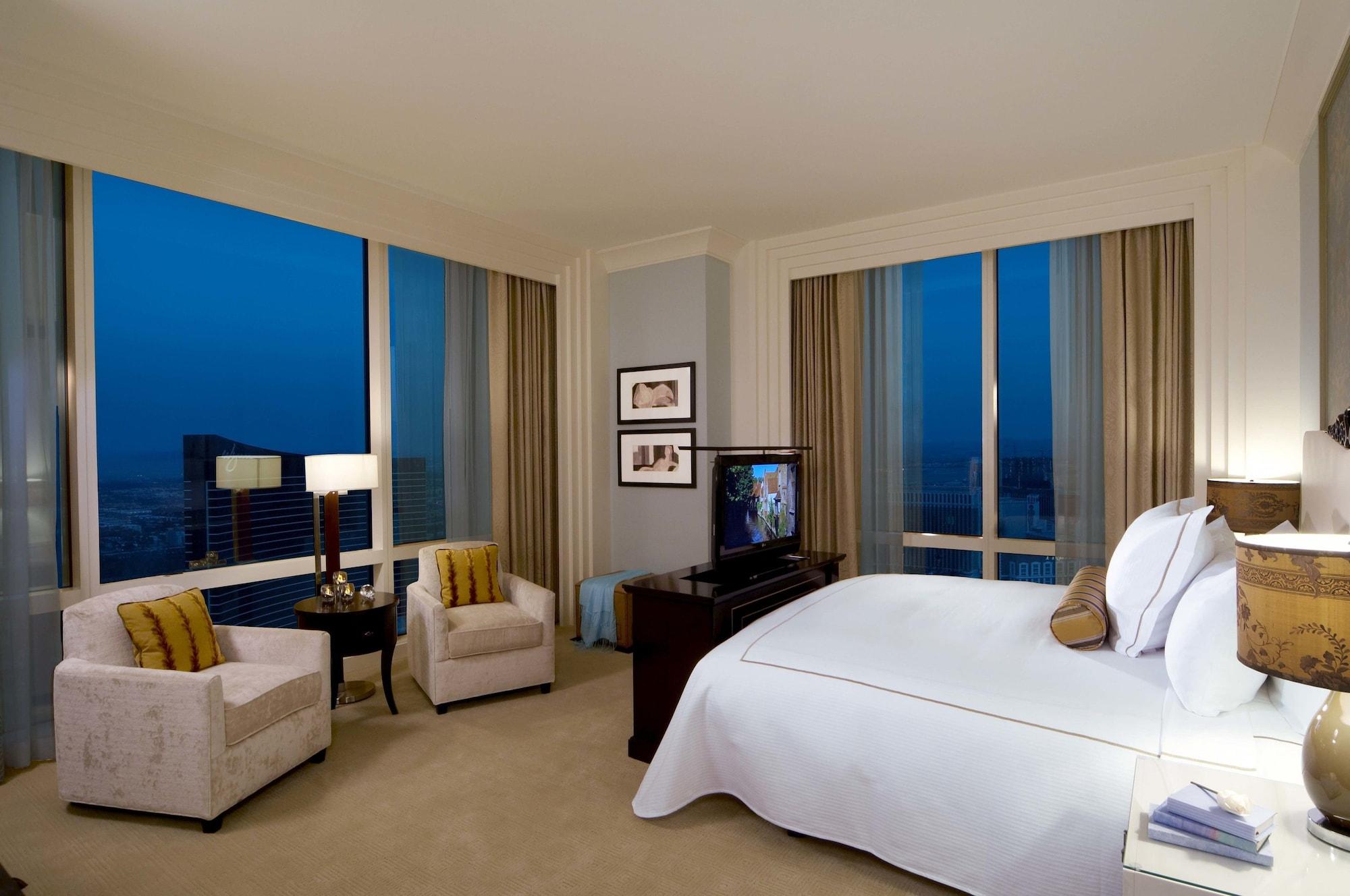 Trump International Hotel Лас Вегас Екстериор снимка The photo shows a modern hotel room featuring a large bed with white linens, positioned near a big window that offers a view of the city. The room has two armchairs with decorative cushions and a small round table beside them. A television is placed 