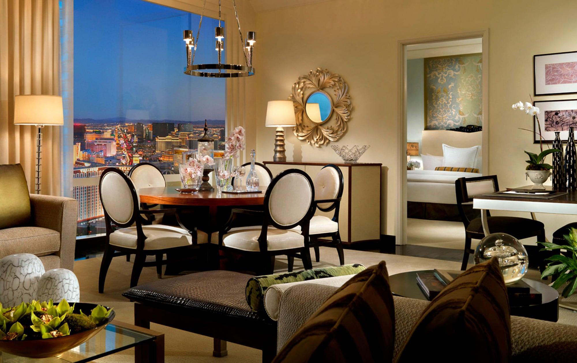 Trump International Hotel Лас Вегас Екстериор снимка The photo shows a luxurious interior space, likely a living room or suite. There is a stylish dining table with elegant chairs positioned near a large window that offers a view of the city skyline, illuminated in the evening. A modern chandelier hang