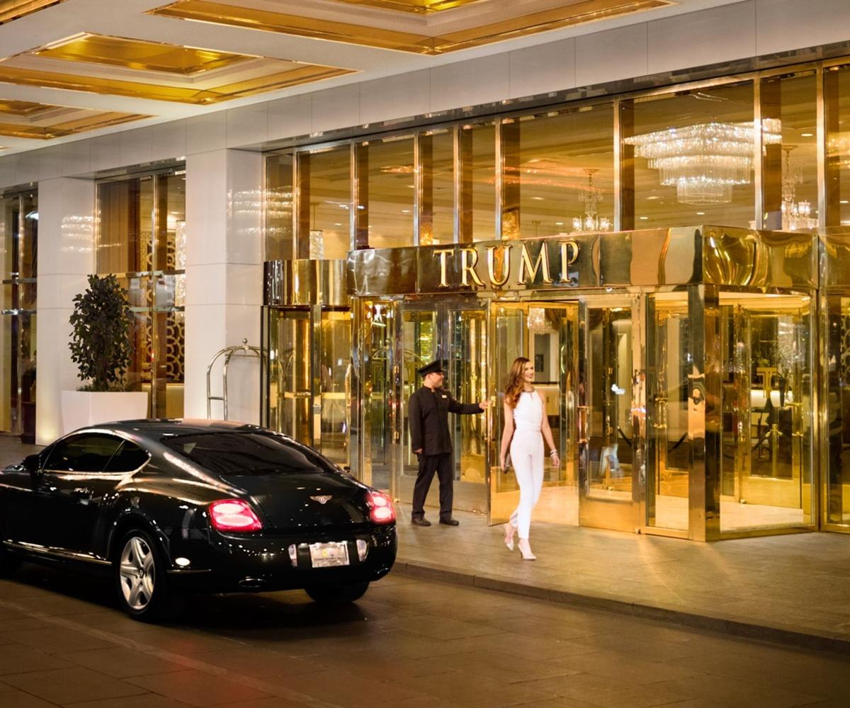 Trump International Hotel Лас Вегас Екстериор снимка The photo shows a luxurious hotel entrance with prominent gold detailing. A well-dressed doorman stands by the door, appearing to assist a woman who is wearing an elegant white outfit as she exits the building. A sleek black car is parked nearby, sug
