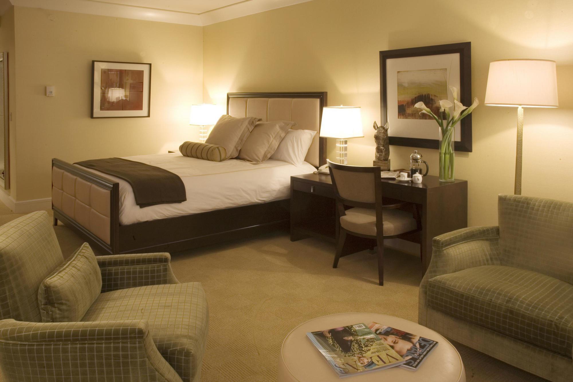 Trump International Hotel Лас Вегас Екстериор снимка The image shows a cozy hotel room with a warm color scheme. The room features a large bed with two decorative pillows, a bedside table with lamps on either side, and a modern desk with a chair. There are two green upholstered armchairs facing a coffe